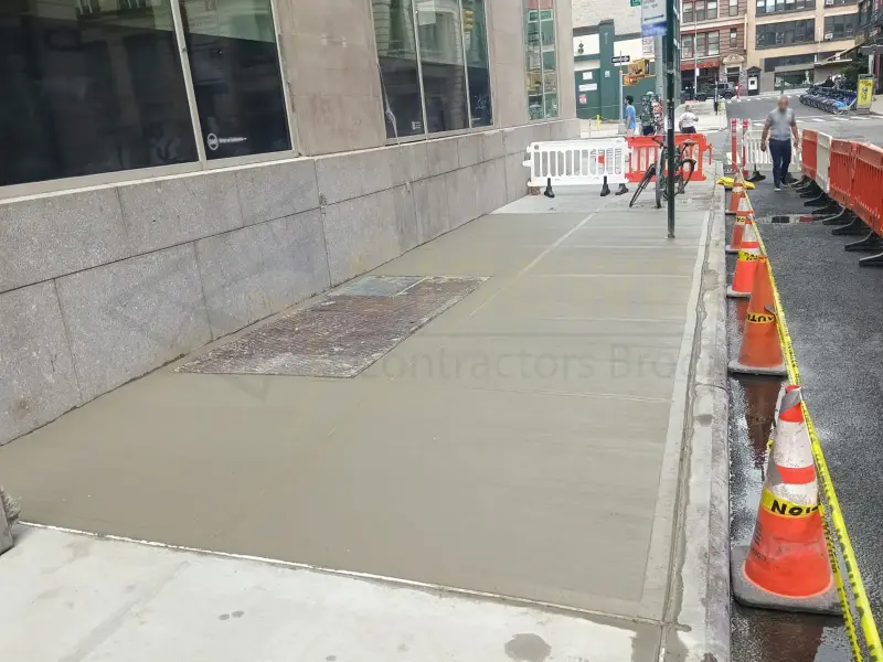 Freshly poured concrete sidewalk beside a commercial building, protected by cones and caution tape